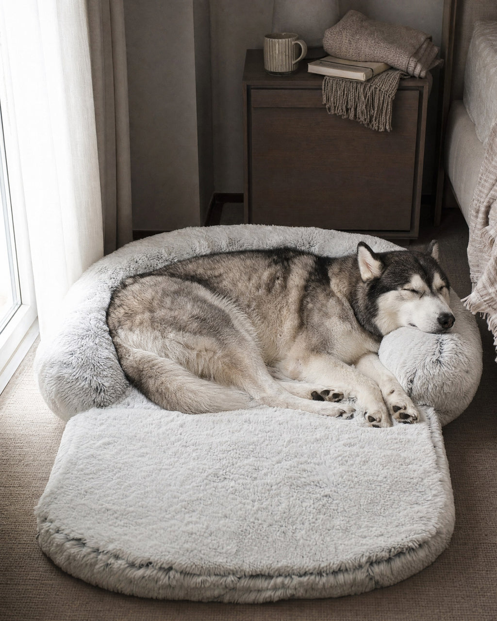Husky resting its head on the rim of a light grey extra-large plush dog bed in a bedroom